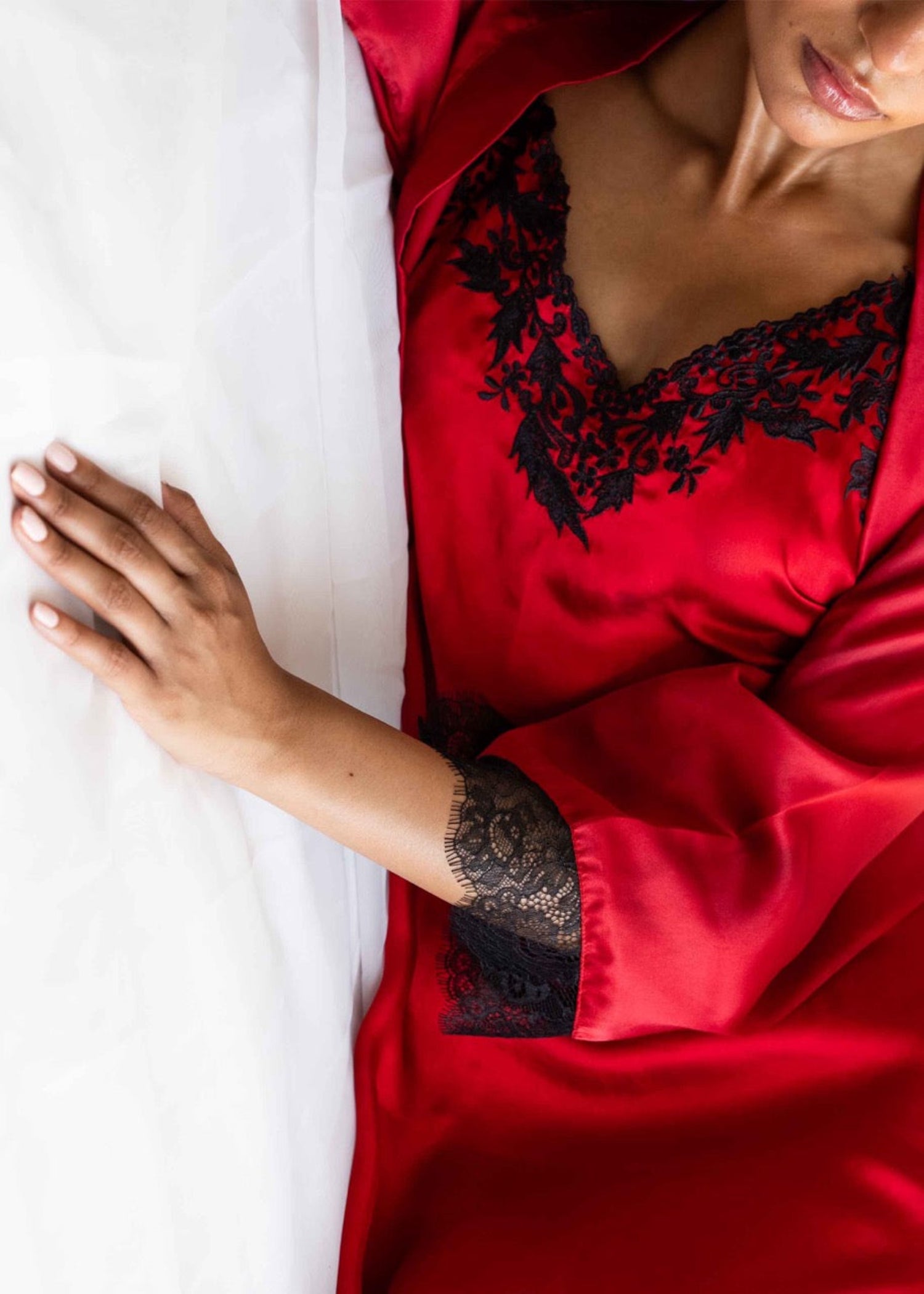 A person lying on a bed wearing a vibrant red silk gown with lace detailing on the neckline and sleeves.