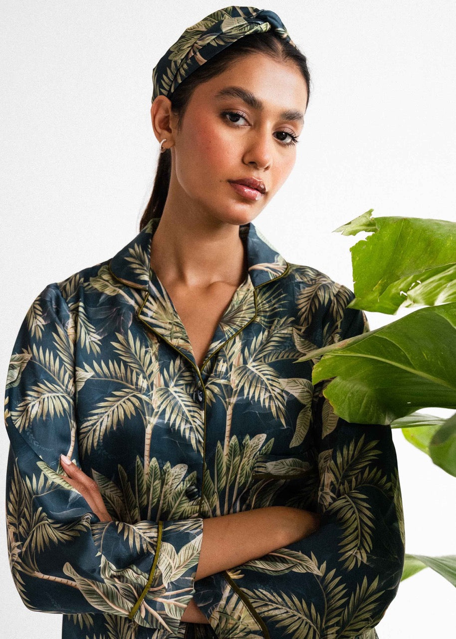 A woman wearing a navy blue and green floral silk co-ord set with a button-up front, standing near a green potted plant.