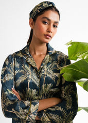 A woman wearing a navy blue and green floral silk co-ord set with a button-up front, standing near a green potted plant.