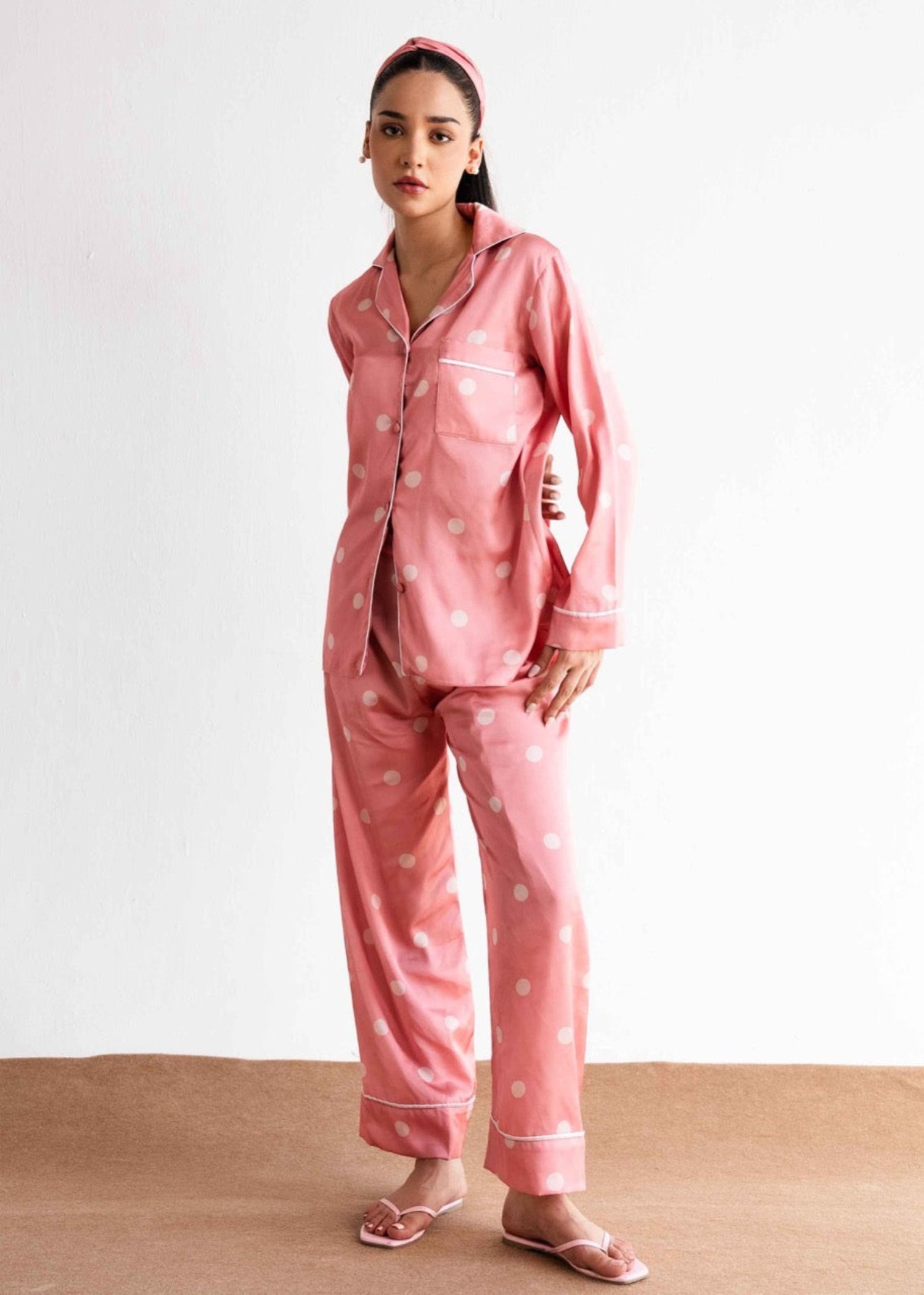 A woman standing and modeling pink sleepwear with white polka dots, consisting of a button-up shirt and pants.