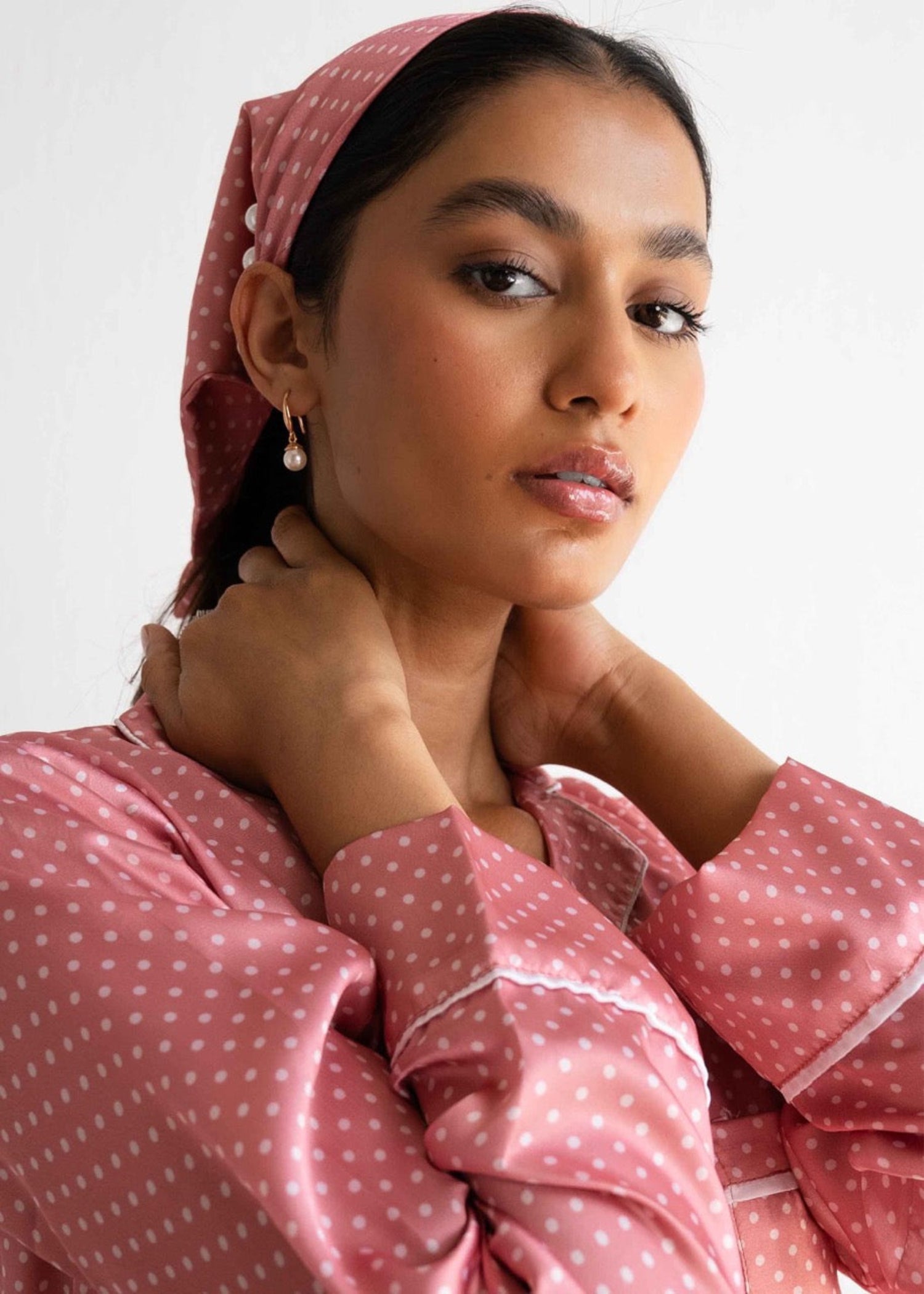 A woman wearing a pink polka dot sleepwear set with a button-up shirt and coordinating pants, posing with her hands behind her head.