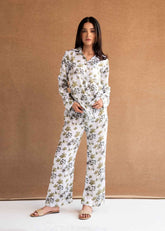 A female model wearing a matching pajama set with a tropical palm tree print on white silk fabric. The set includes a button-down shirt and coordinating pants.