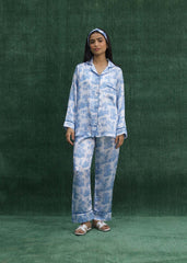 A woman standing against a green background, wearing a white silk pajama set with powder blue leaf prints, consisting of a button-down shirt with front pockets and trousers with lined hems.