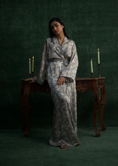 A model wearing a light gray silk gown with floral prints and a matching waist belt, standing against a green background. - Larosa