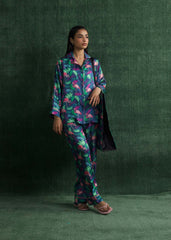 A woman standing against a green backdrop, wearing a navy blue silk pajama set with vibrant multicolor prints and pink flamingo designs. The outfit includes a button-down shirt and front pockets.