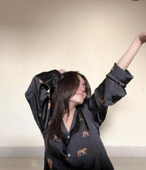 A woman wearing a black silk sleep set with leopard print patterns, including pants and a top, posing with one hand raised and an expression of joy.